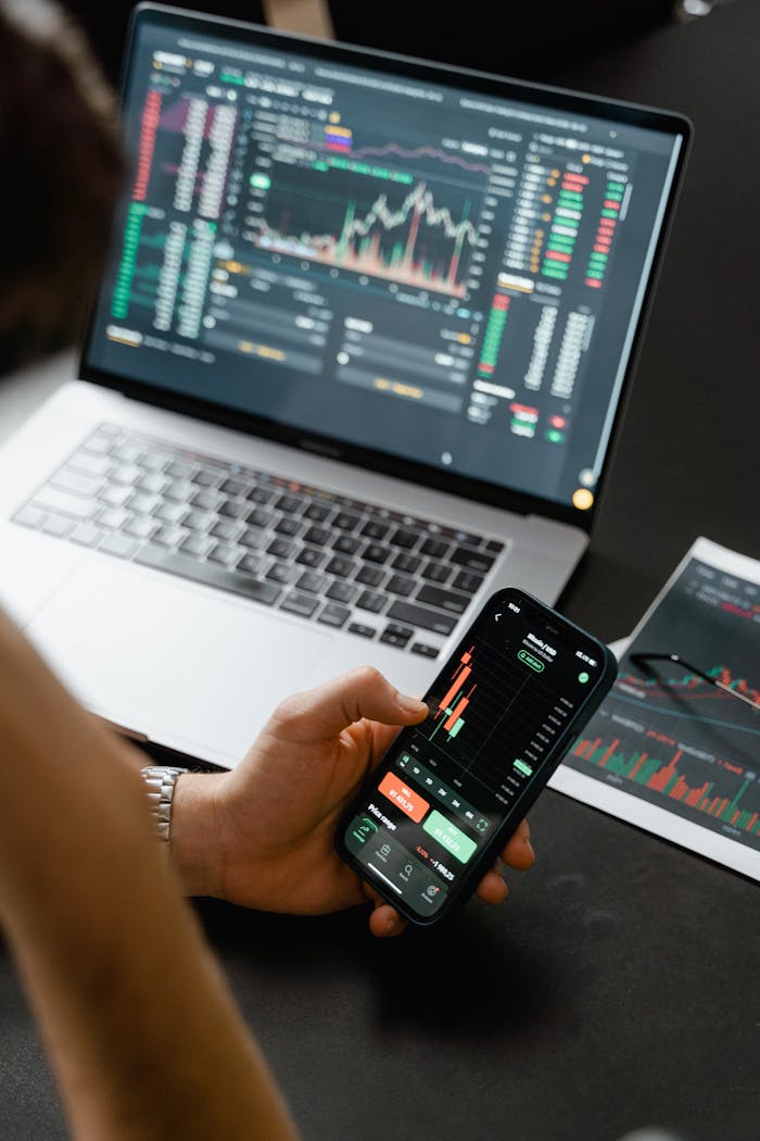 portfolio-04 Person analyzing stock market data on a smartphone with a laptop displaying financial graphs in the background.